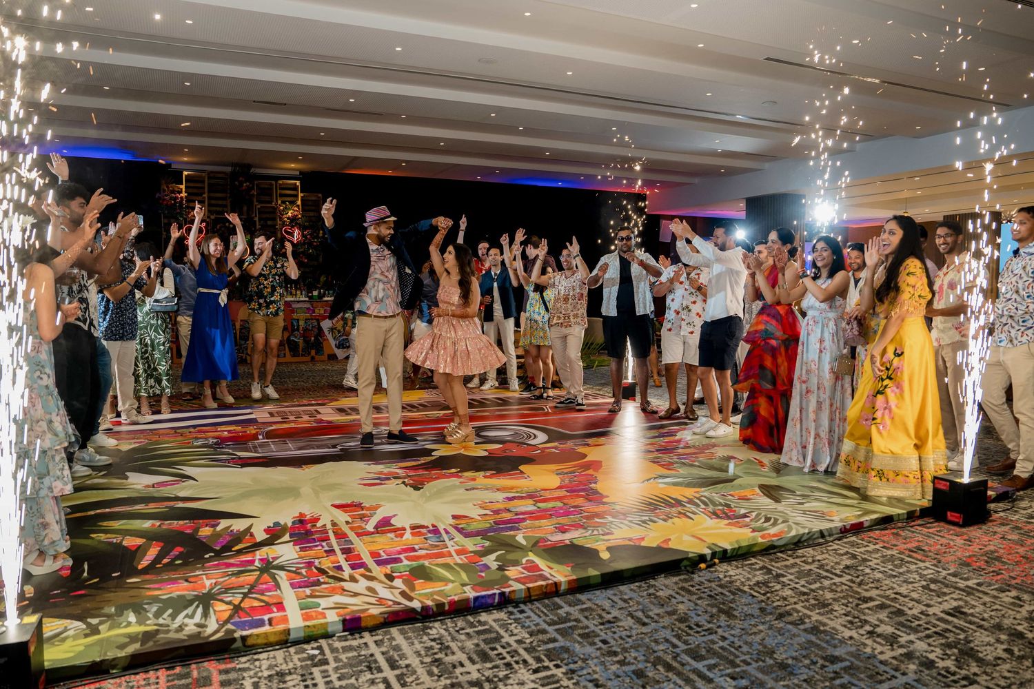 Wedding guests dance and celebrate on a colorful patterned dance floor at an indoor reception venue.