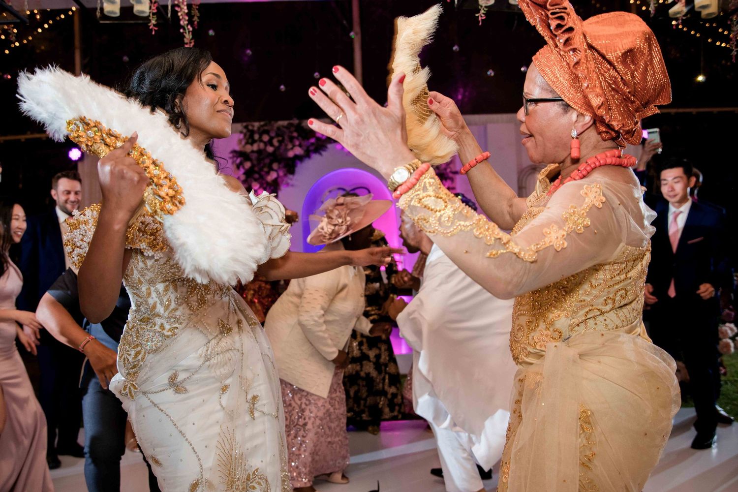 Wedding guests dancing enthusiastically at a reception with decorative lighting and floral arrangements.