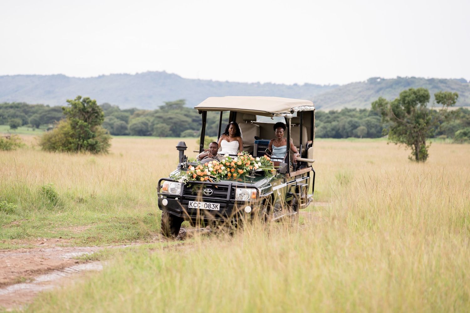 Safari vehicle drives through tall grass in the African savanna with hills in background.