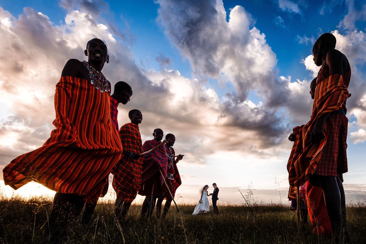 Dramatic sunset silhouettes of people in traditional Maasai clothing standing on savanna grassland.