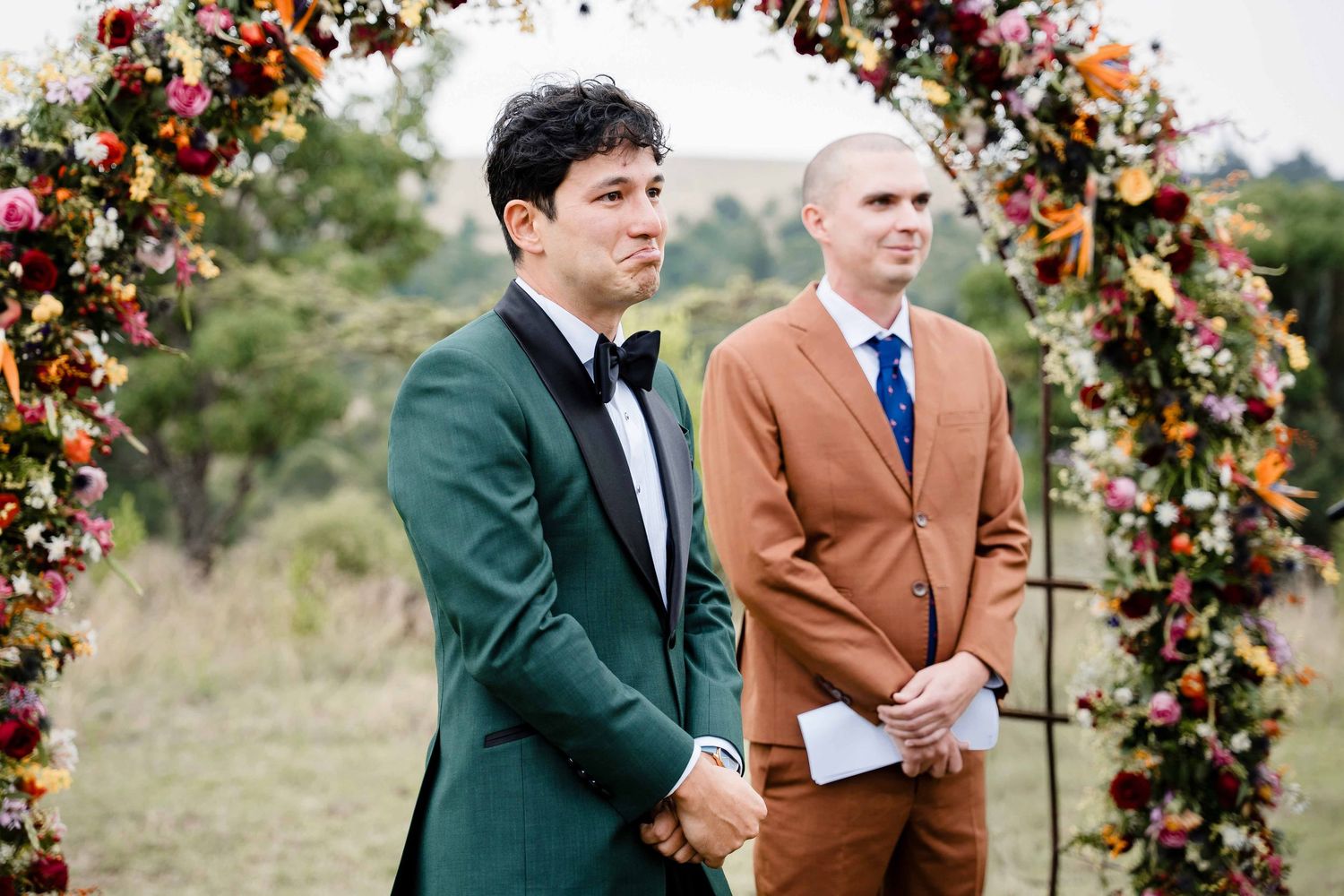 Two men in formal attire stand together under a floral arch at an outdoor wedding ceremony.