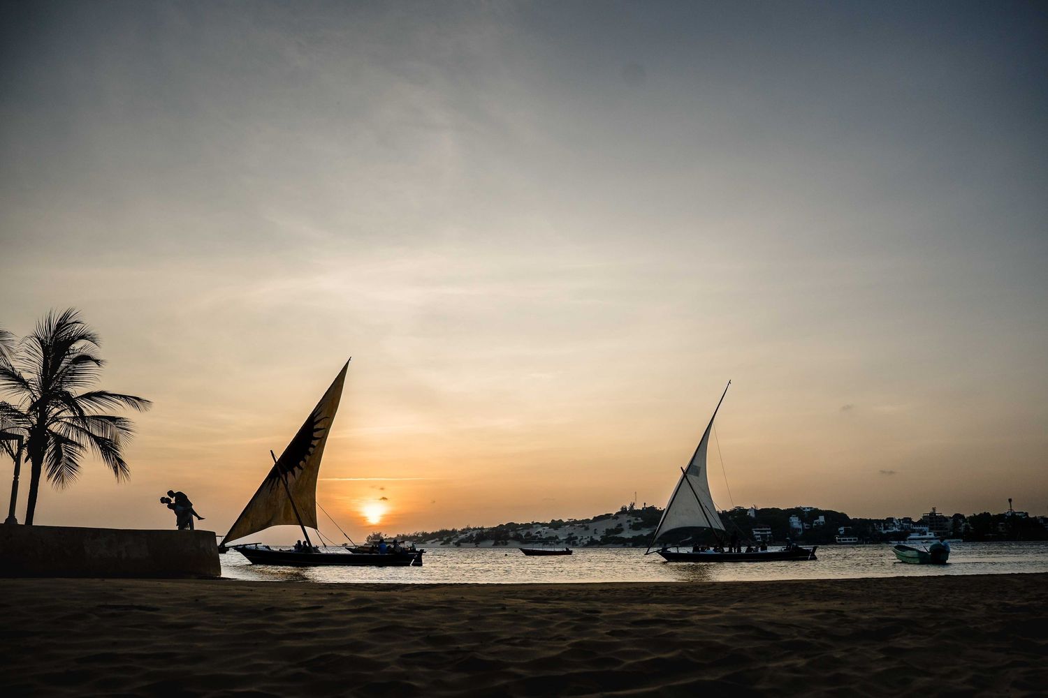 Silhouettes of palm trees and sailboats against a vibrant sunset over a tropical beach.