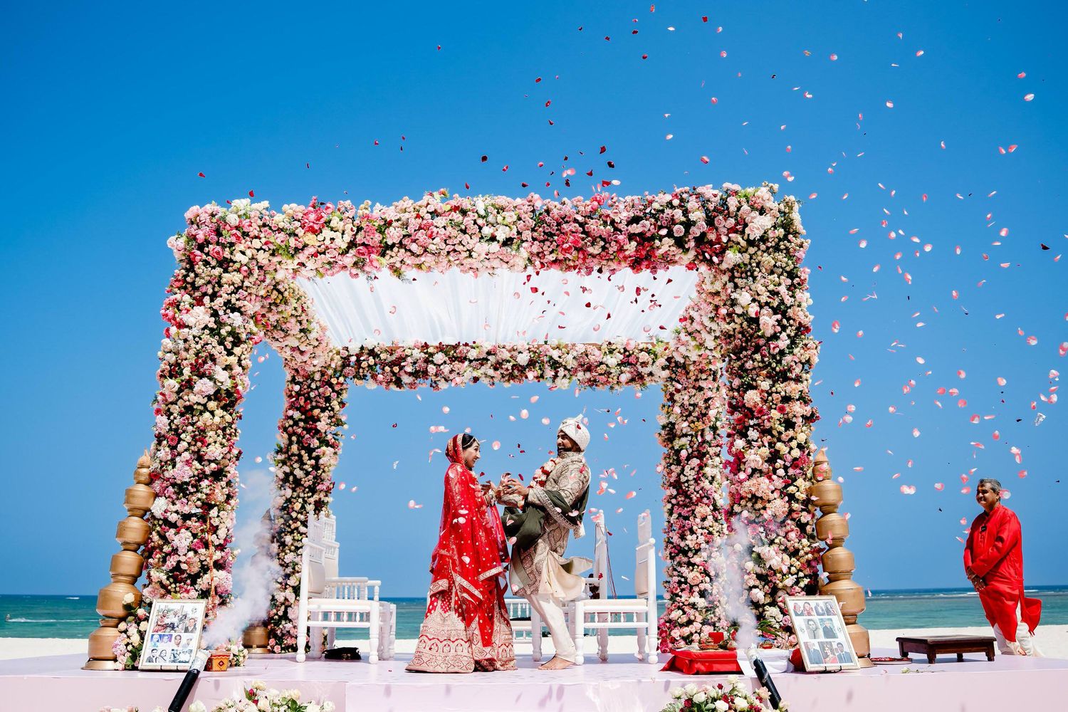 Pink flower petals rain down during a beach wedding ceremony under a floral arch against bright blue sky and ocean.