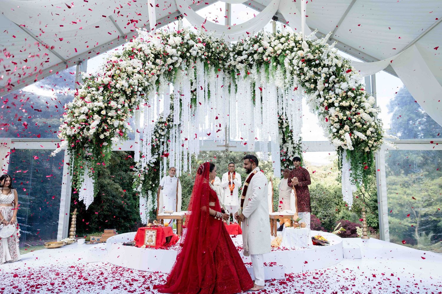 Wedding ceremony under a white floral arch with red rose petals falling.