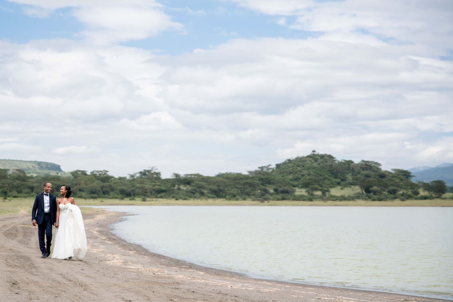 A wedding couple walks along a serene lakeshore with mountains in the distance.