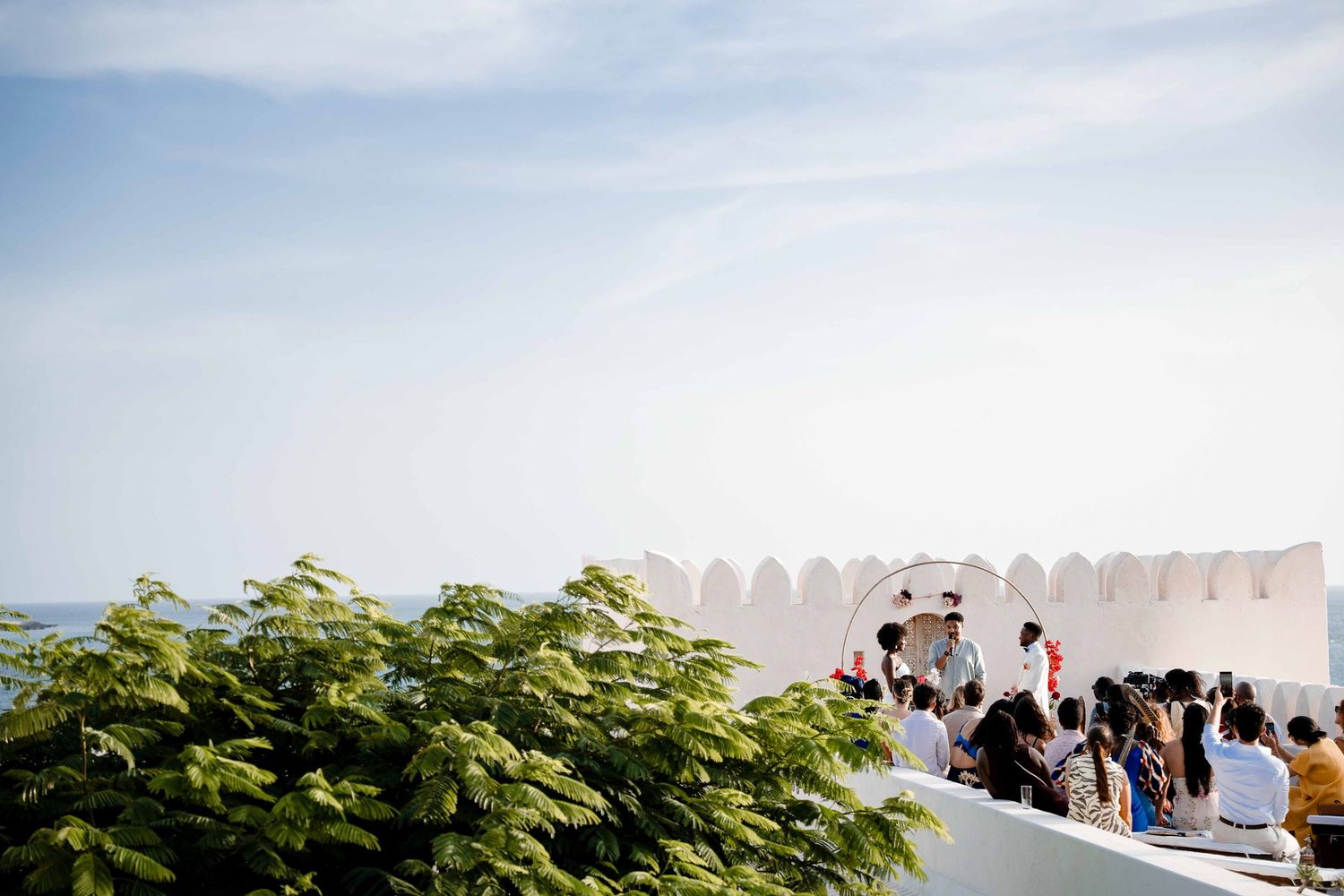 A white stone balcony overlooking the ocean with green foliage and people gathered at a coastal venue.