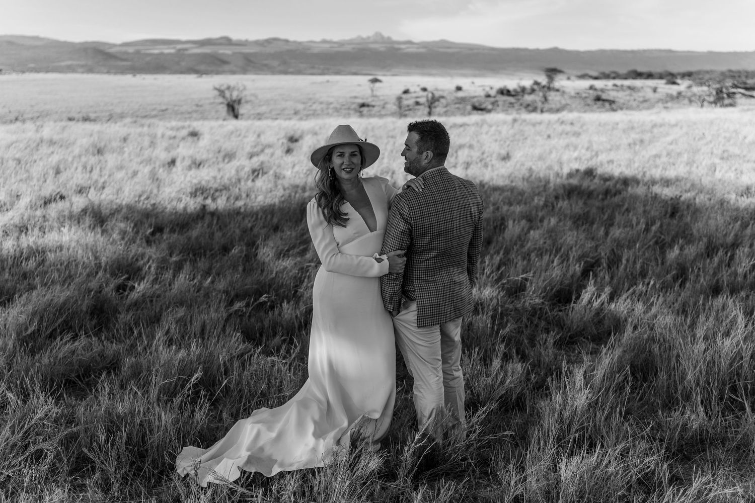 Black and white photo of a couple standing in tall grass with mountains in distance.
