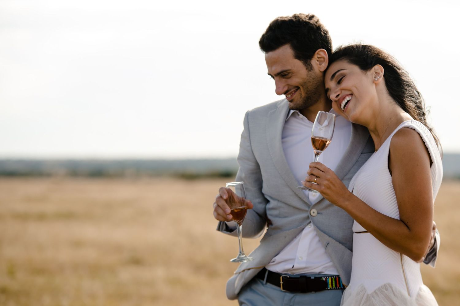 A couple laughs together while sharing wine in a sunlit field.