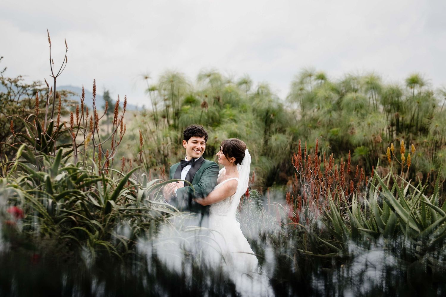 A couple embraces in a romantic outdoor garden setting with red flowers and greenery.