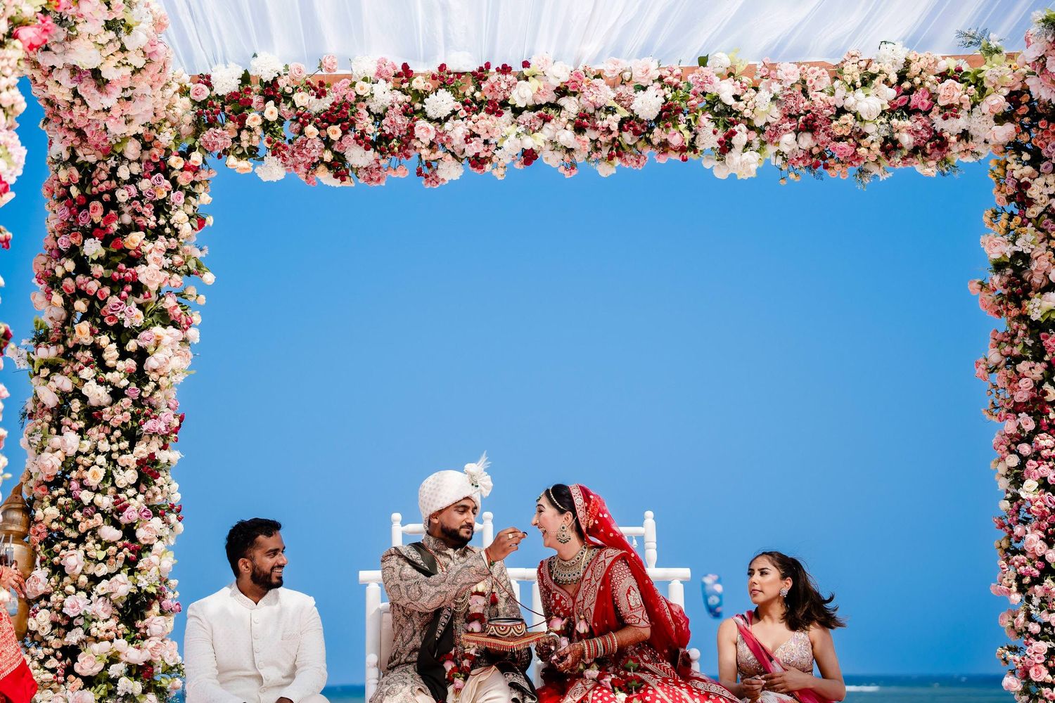 Couple celebrates during traditional Indian wedding ceremony under floral archway with blue sky backdrop.