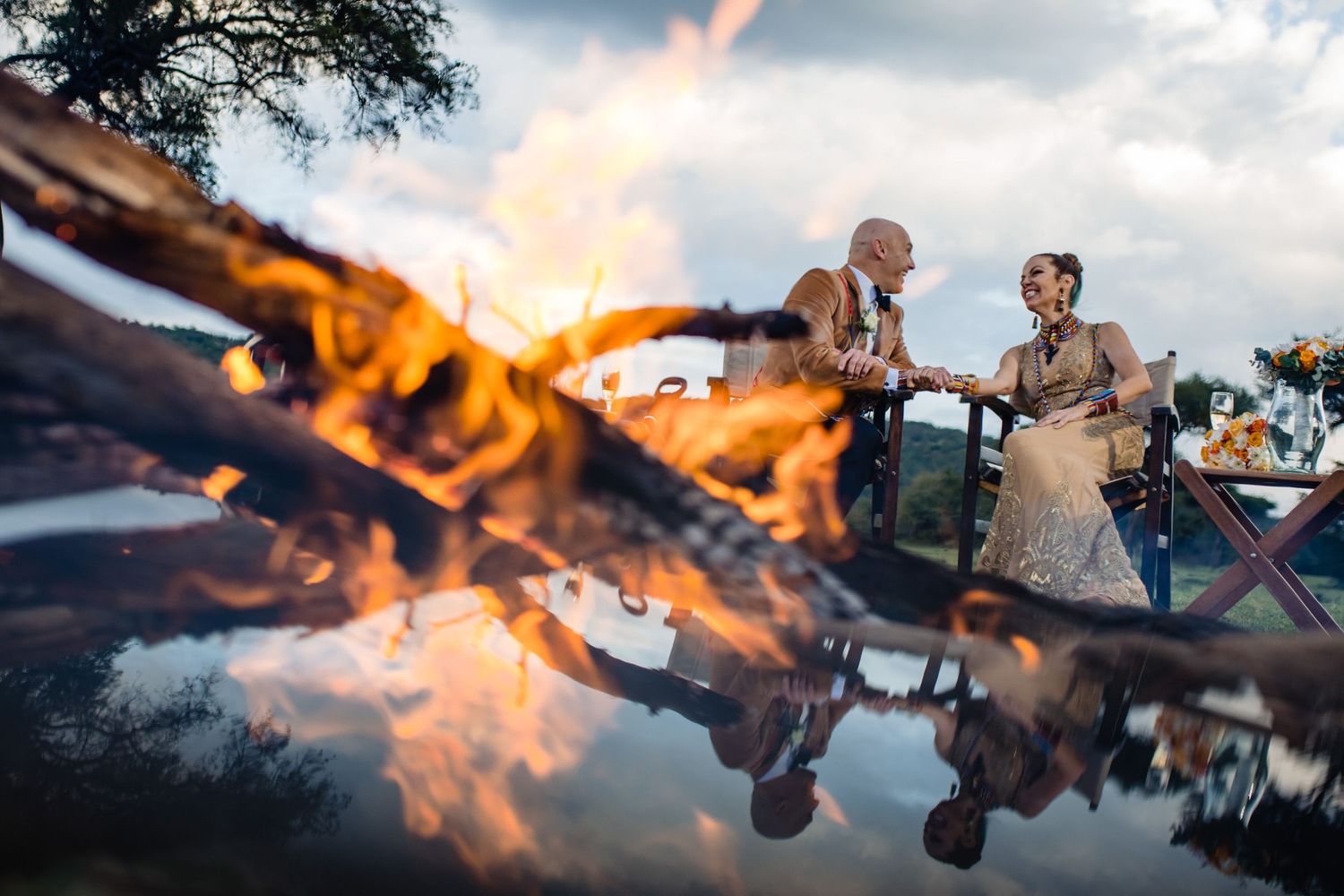 Couple by a bonfire with reflection in water during sunset.