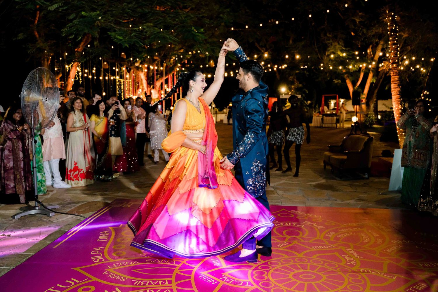 Colorful Indian wedding celebration with guests dancing under string lights at an outdoor evening reception.