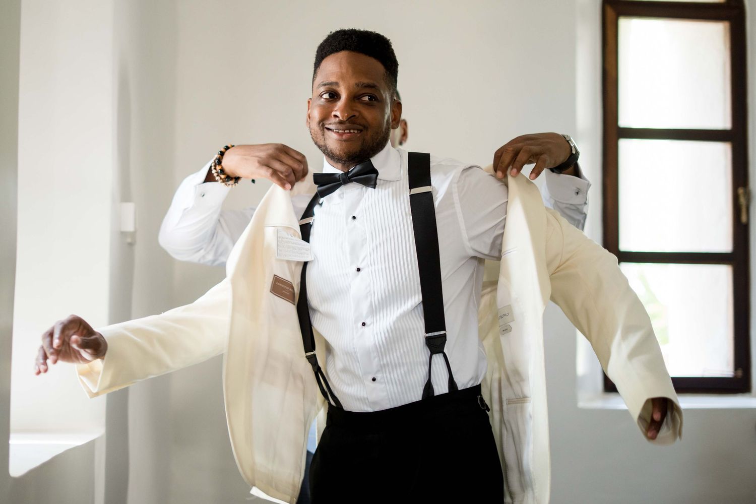 A person in a white tuxedo jacket and black suspenders poses energetically against a bright interior wall.
