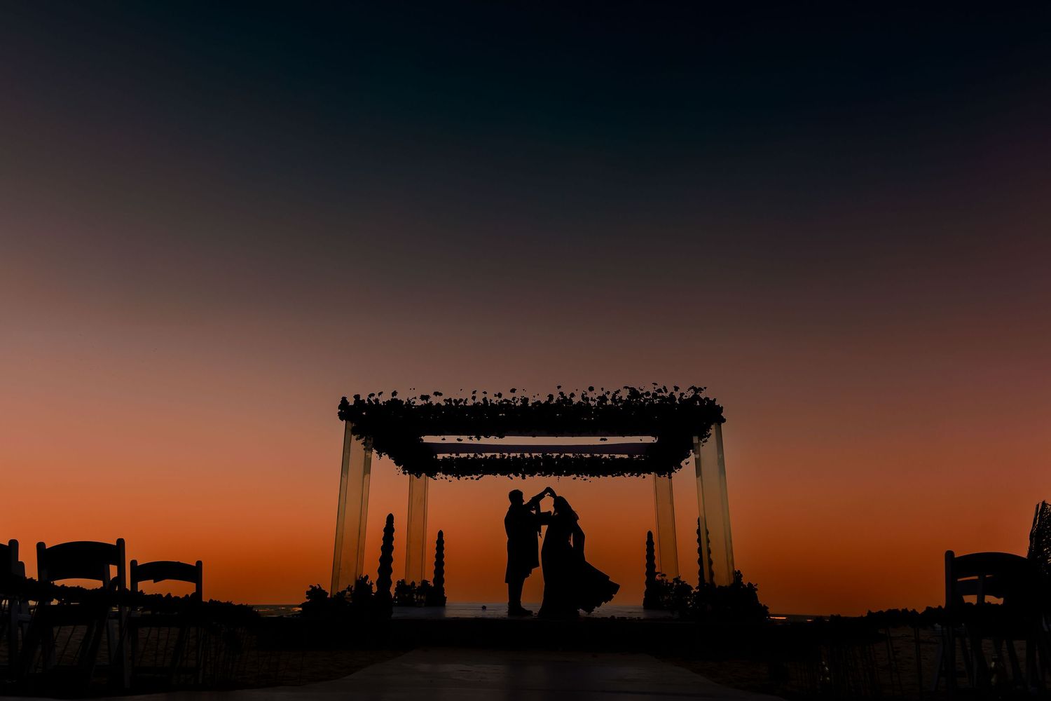 A romantic outdoor wedding ceremony silhouetted against a dramatic orange and purple sunset sky.
