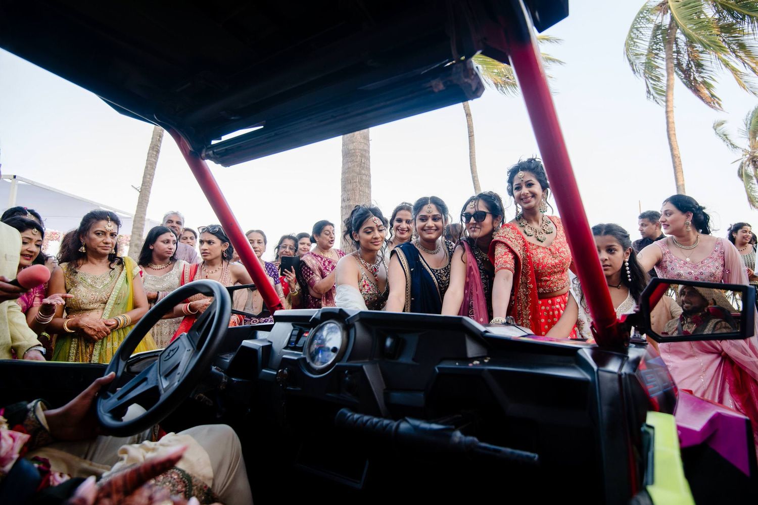 A crowd of enthusiastic people gather around a car at a tropical beach event with palm trees in the background.