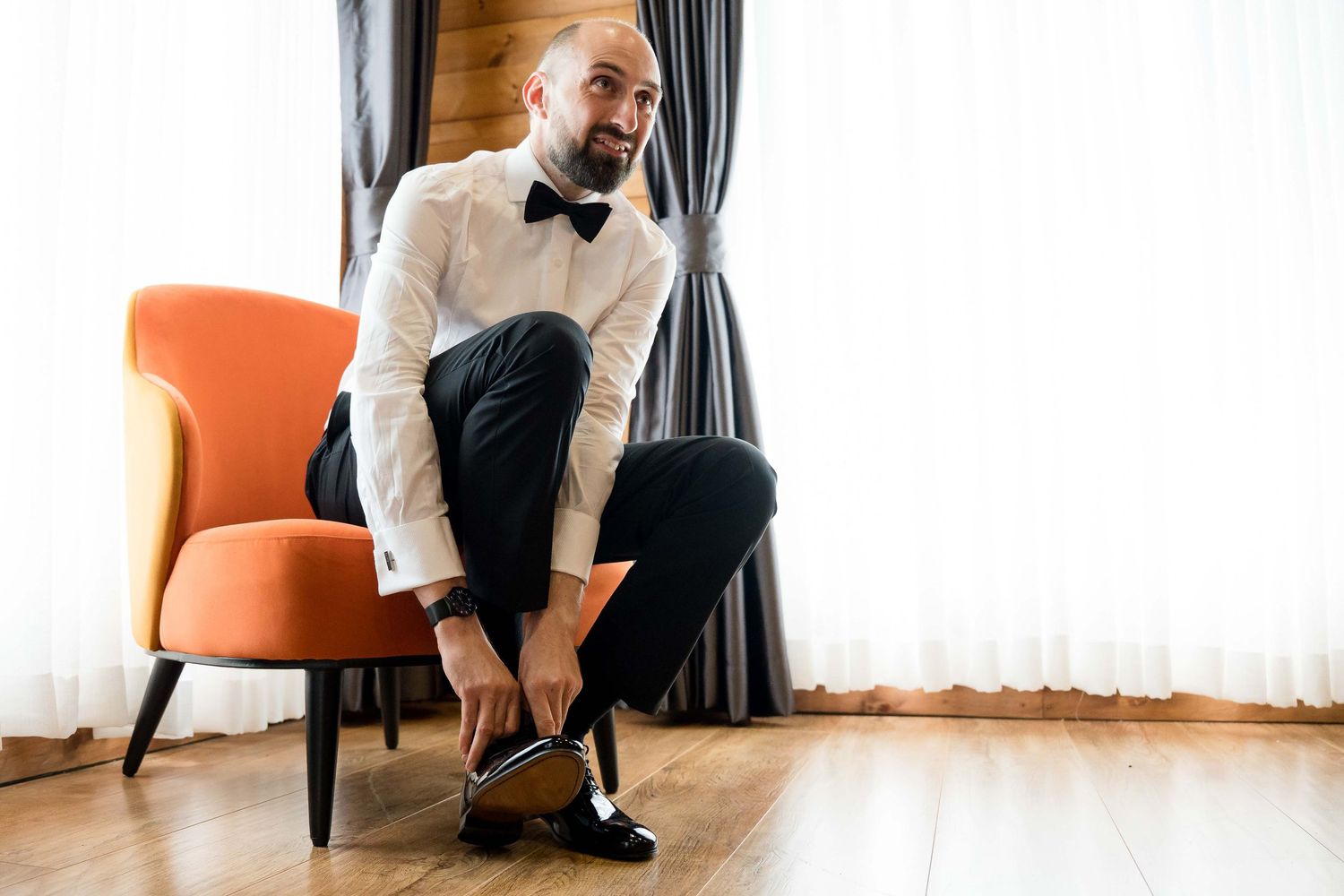 Someone in a black suit and bow tie sits elegantly in an orange armchair near curtained windows.