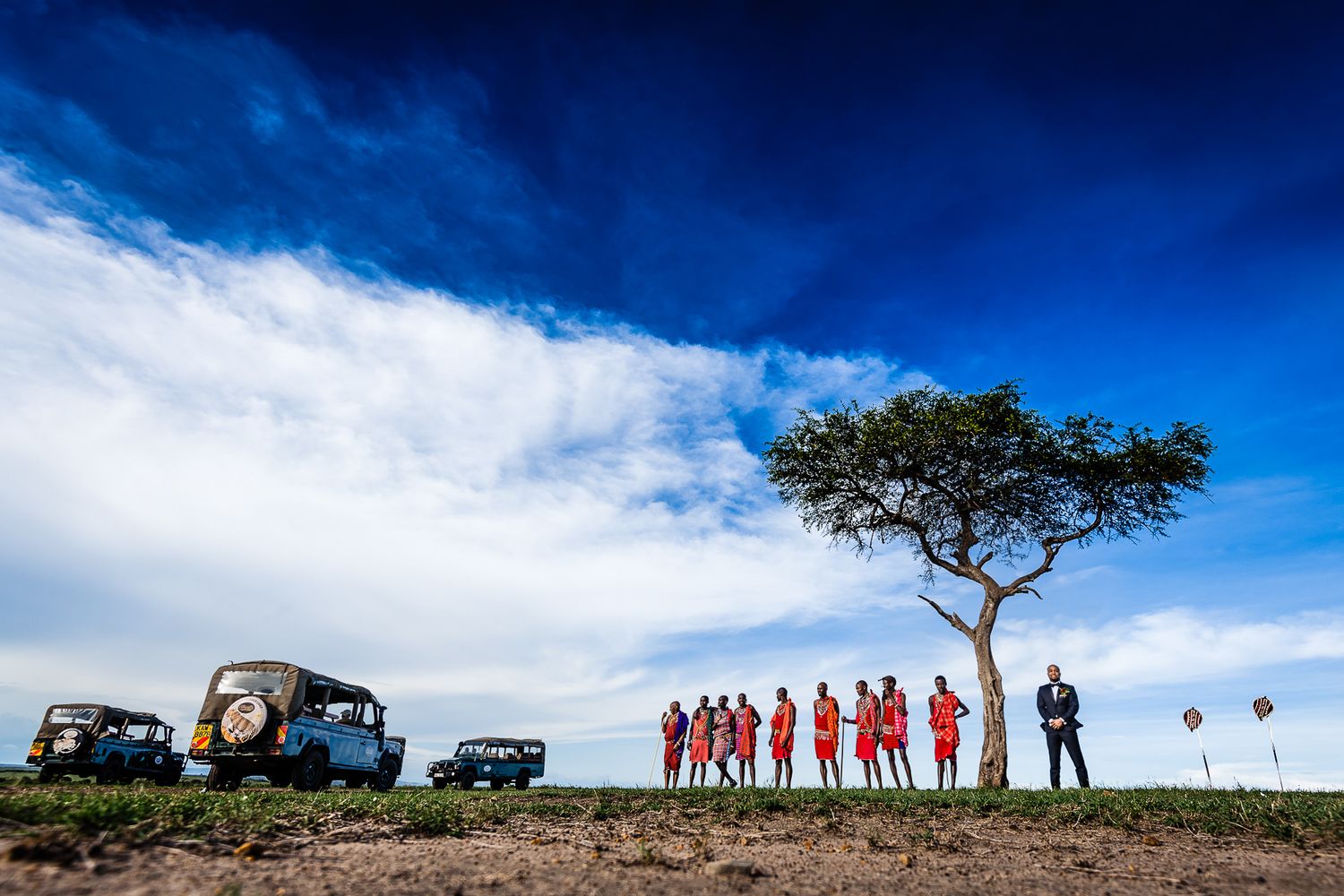 Safari vehicles parked under lone acacia tree with group silhouetted against dramatic sky.