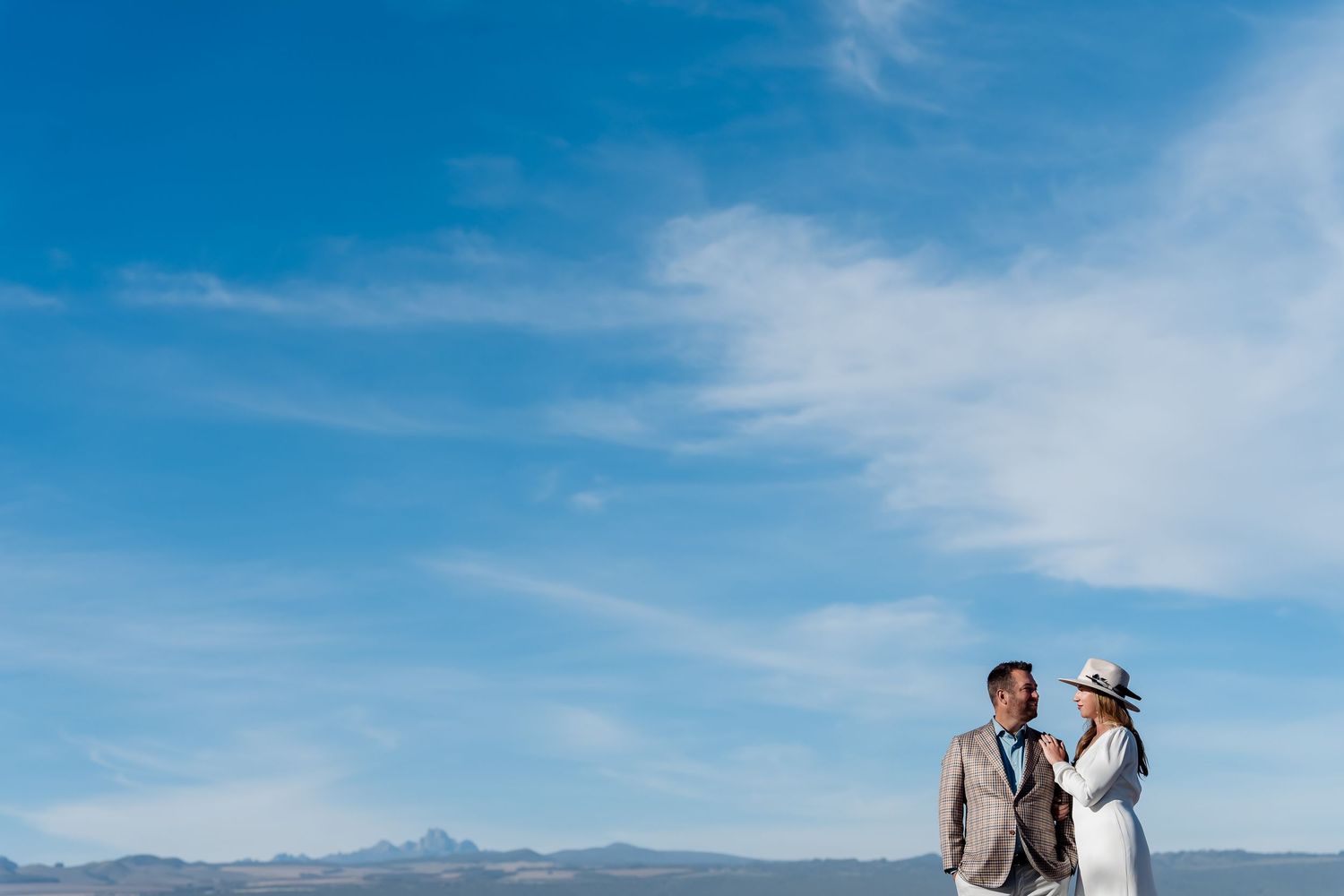 A couple in formal attire stands together against a bright blue sky and mountain backdrop.