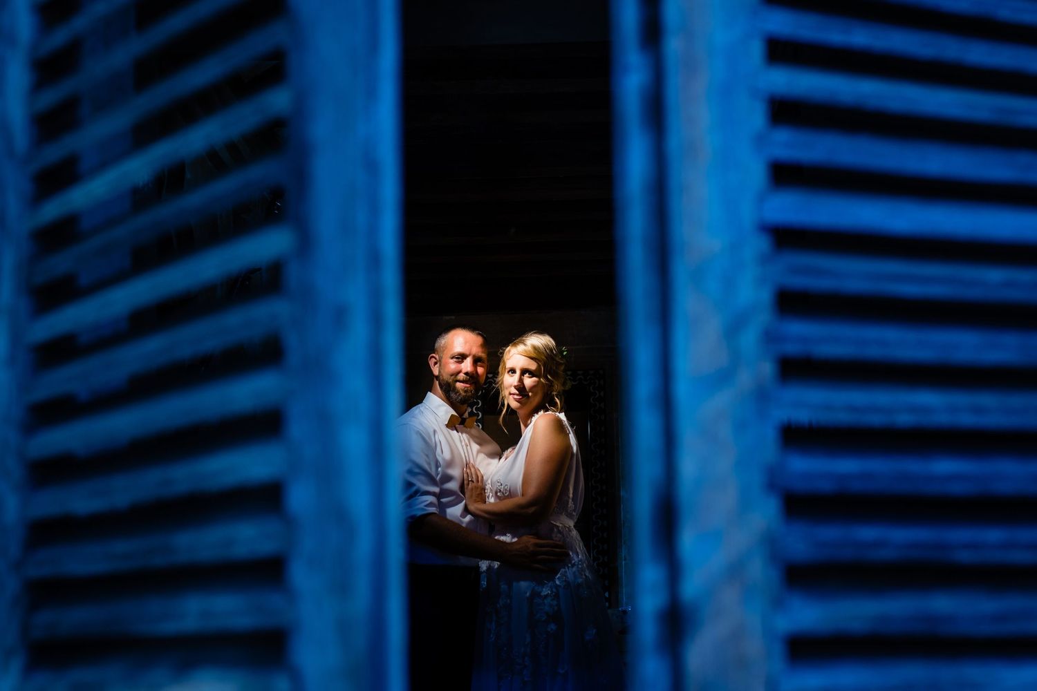 Intimate portrait through blue shutters showing couple in romantic embrace.