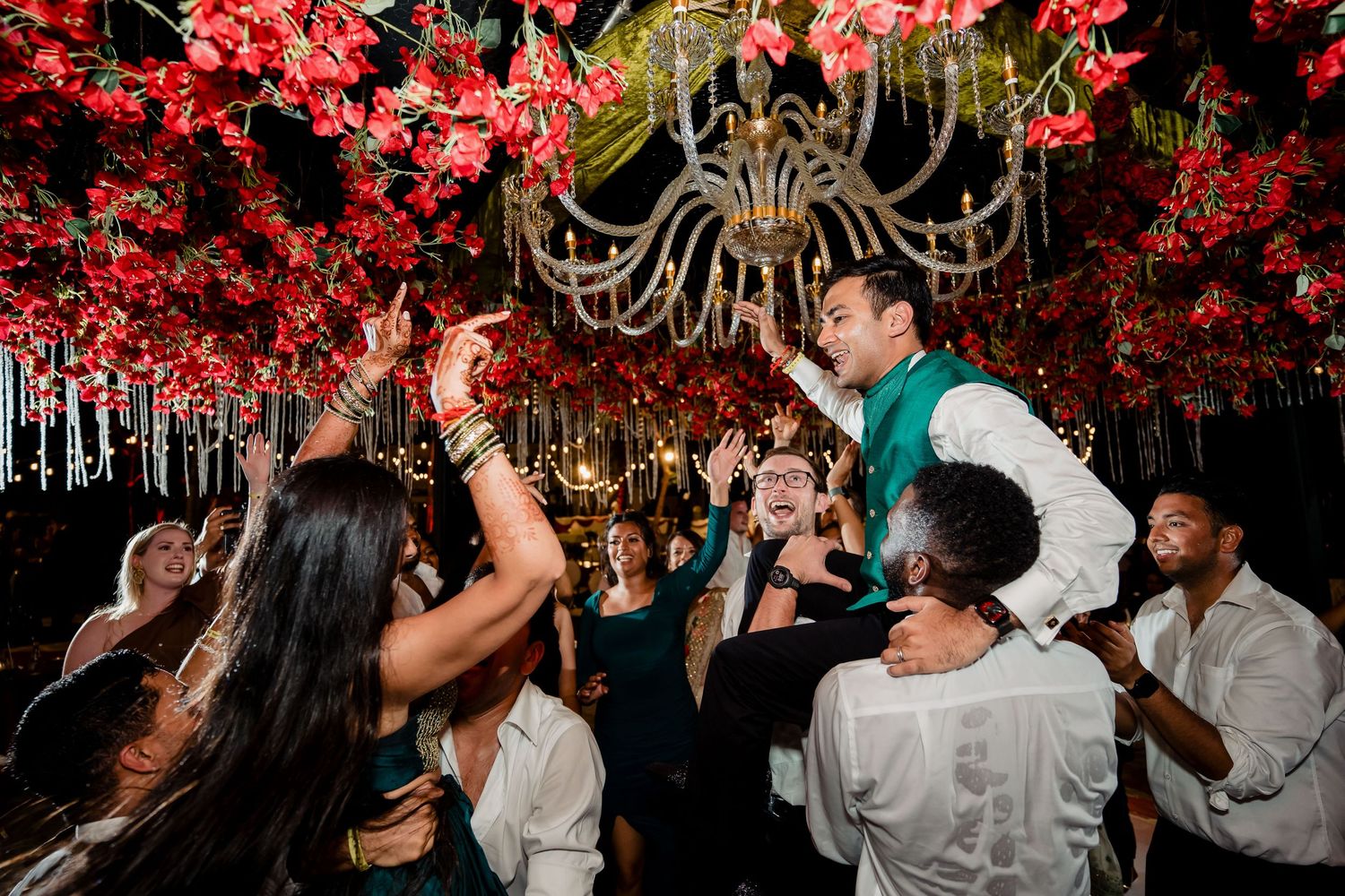 Wedding guests dancing and celebrating under red floral ceiling decorations and chandelier.