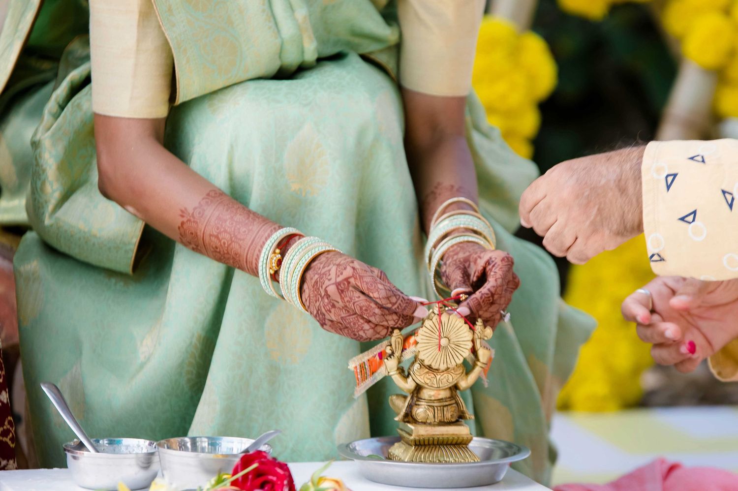 A pair of henna-adorned hands perform a traditional Indian religious ceremony with a small golden statue.
