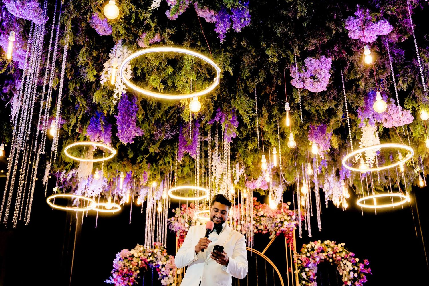 A romantic wedding reception venue decorated with hanging purple flowers and glowing string lights creating a magical atmosphere.