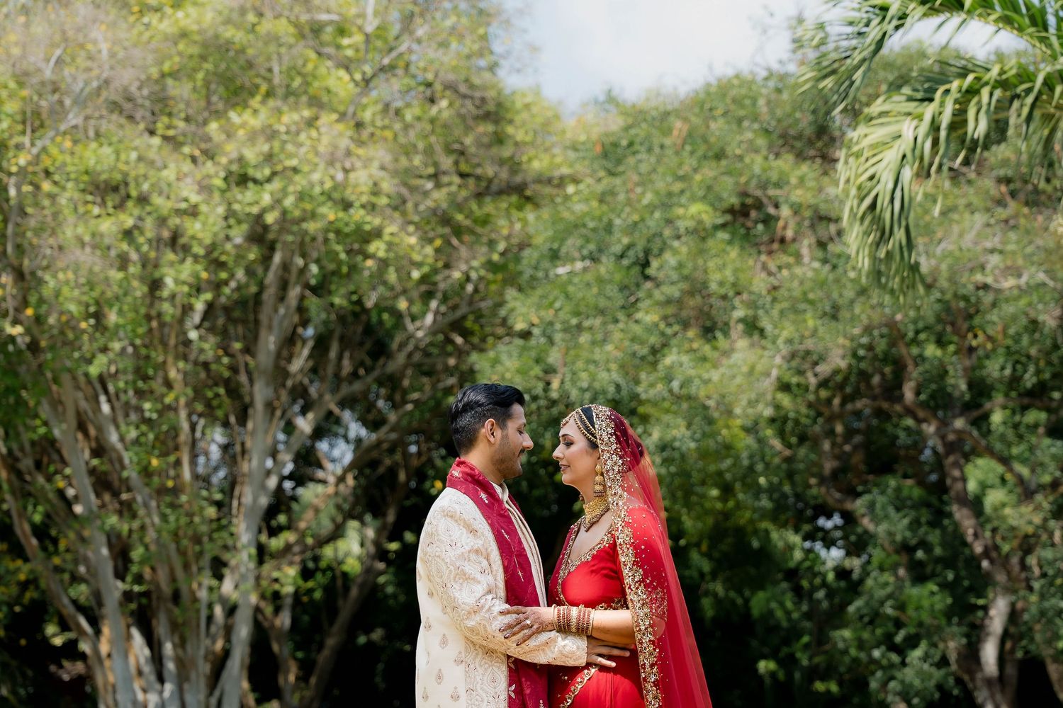 A couple in traditional South Asian wedding attire stands together in a lush garden setting.