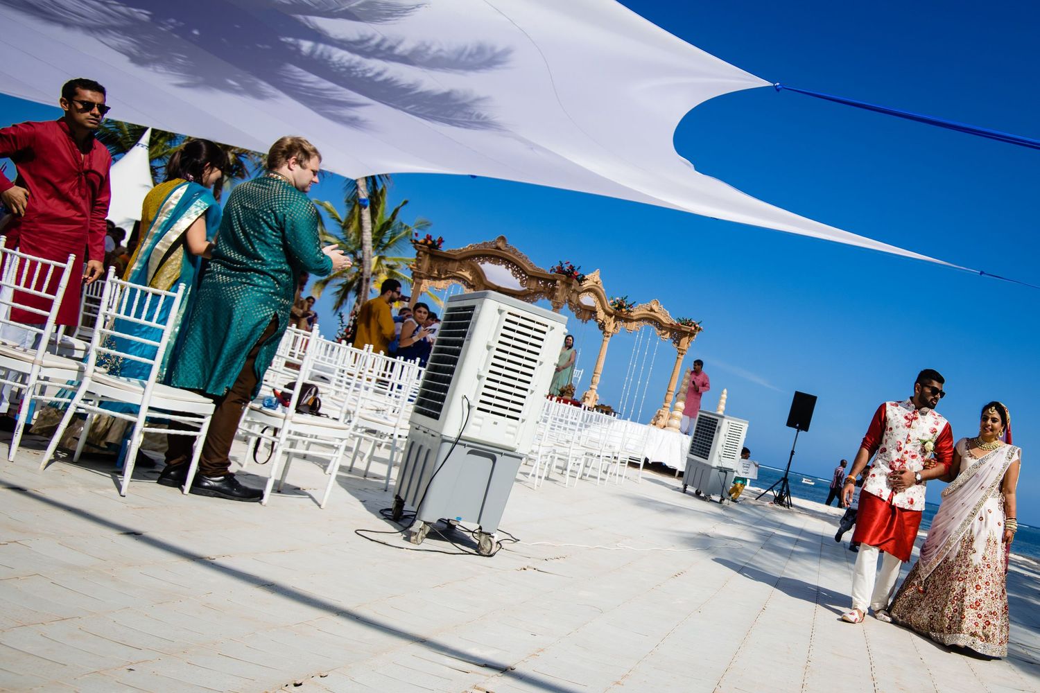 Outdoor beach wedding setup with white chairs and fabric canopy against blue sky.