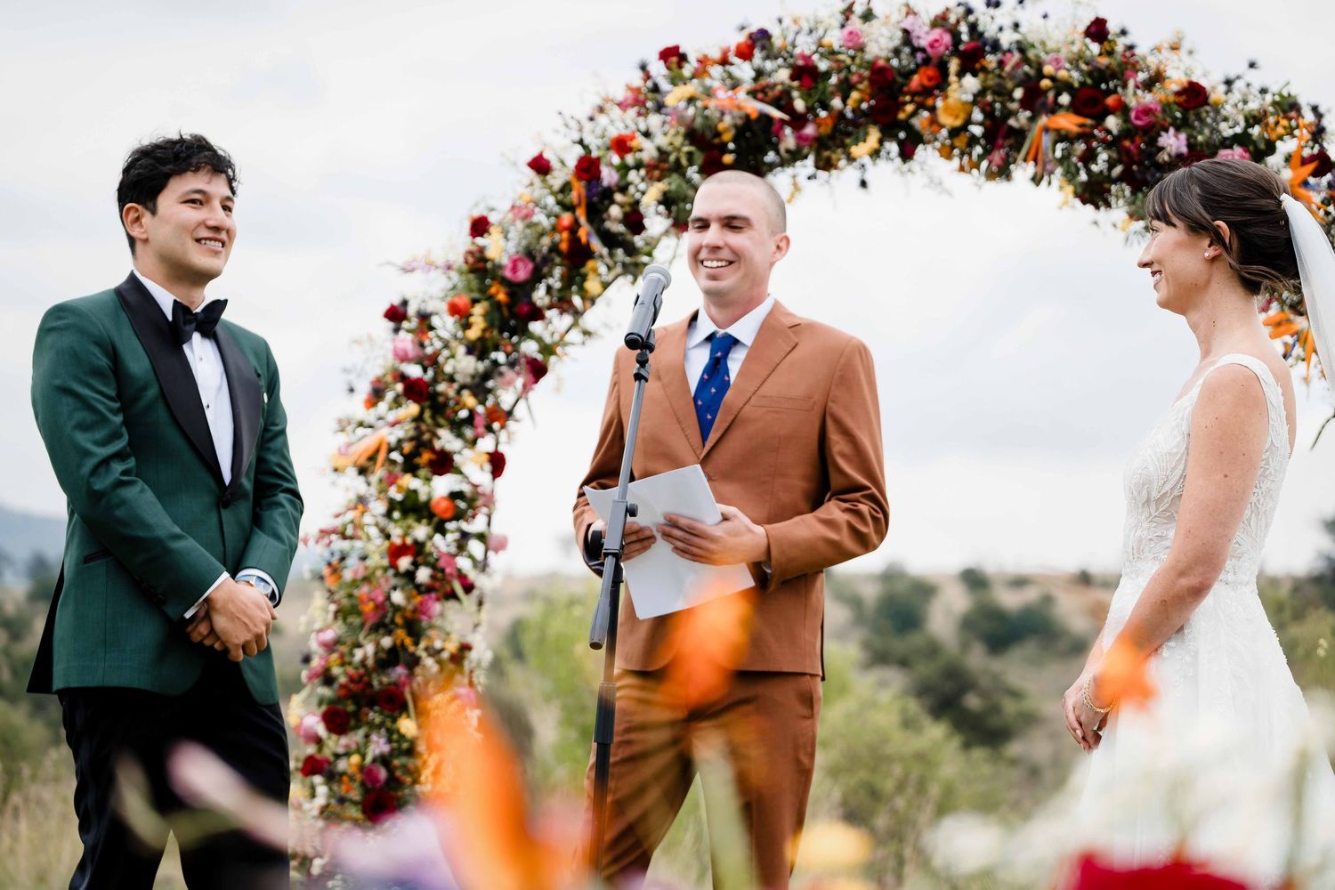 A wedding ceremony takes place outdoors under a floral arch with mountains in the background.
