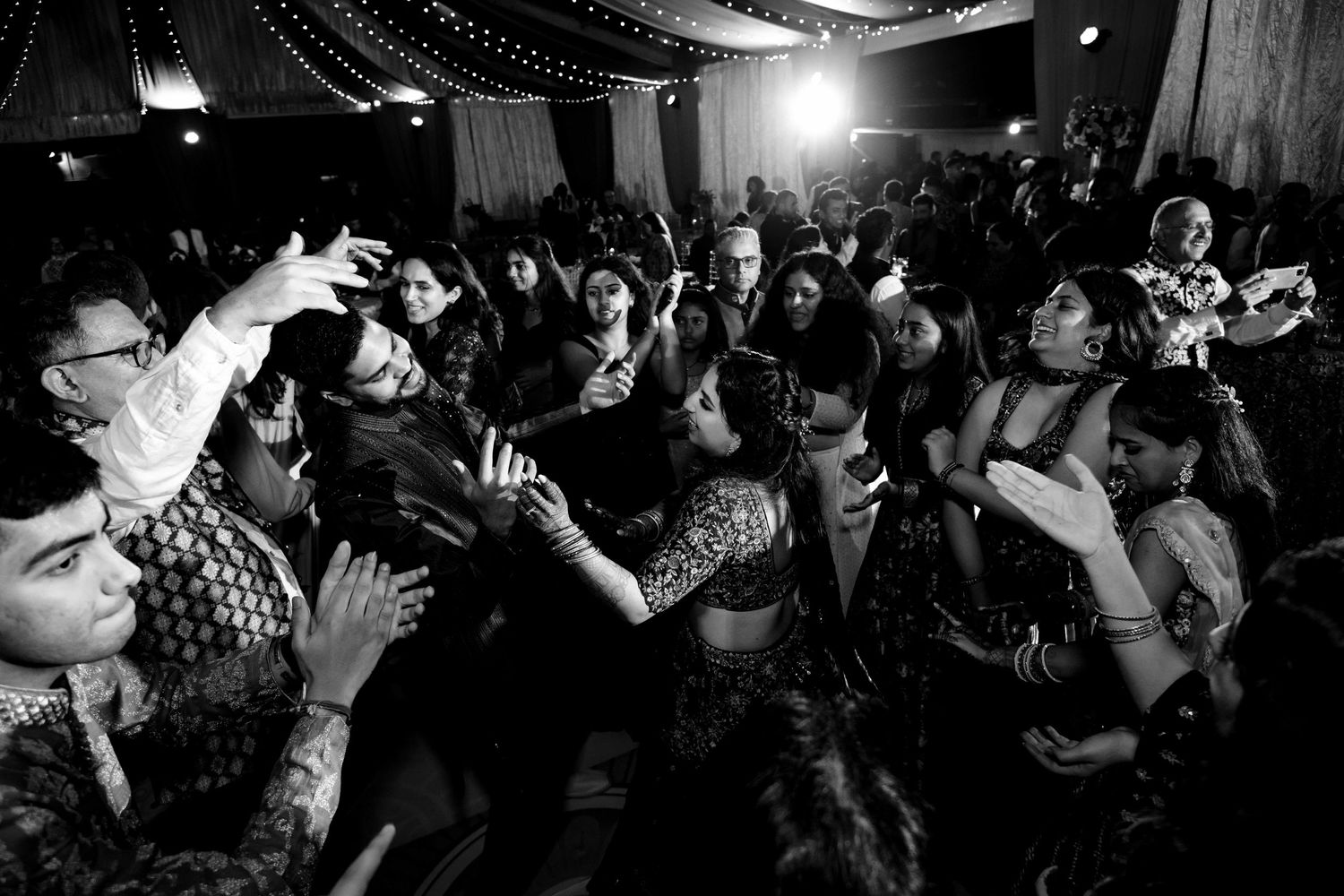 A crowd dances energetically at an Indian celebration captured in black and white photography.