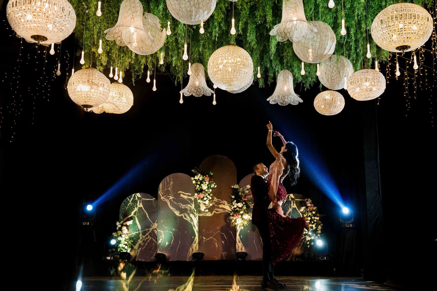 White paper lanterns hang from a ceiling with greenery and blue lighting creating an enchanting party atmosphere.