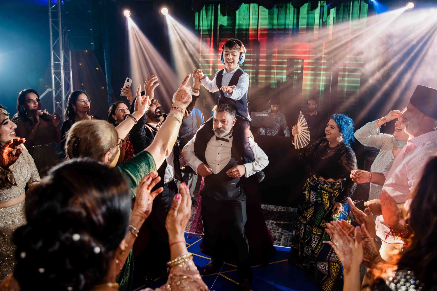 Enthusiastic crowd lifts a guest at a lively wedding reception with dramatic stage lighting and atmosphere.