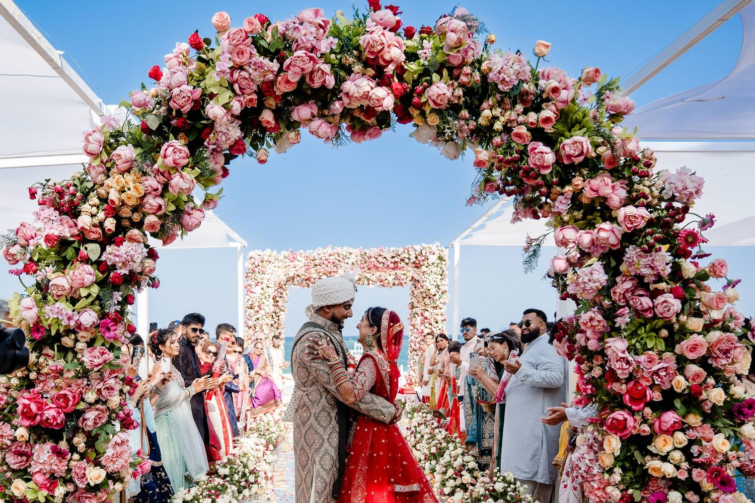 A wedding ceremony setup with a stunning pink and white floral arch against a bright blue sky.