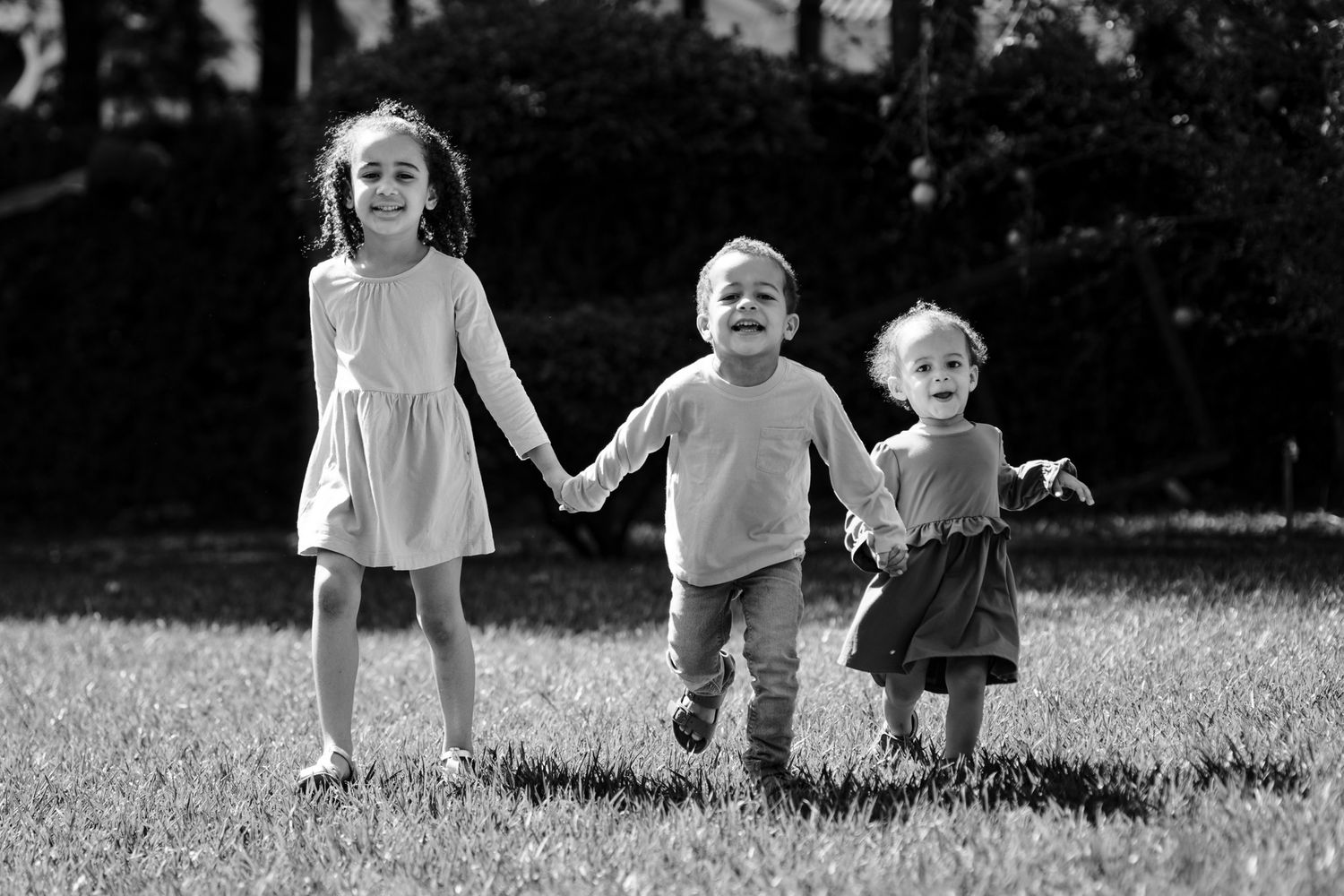 Three young children hold hands while standing together in a grassy field in black and white photography.