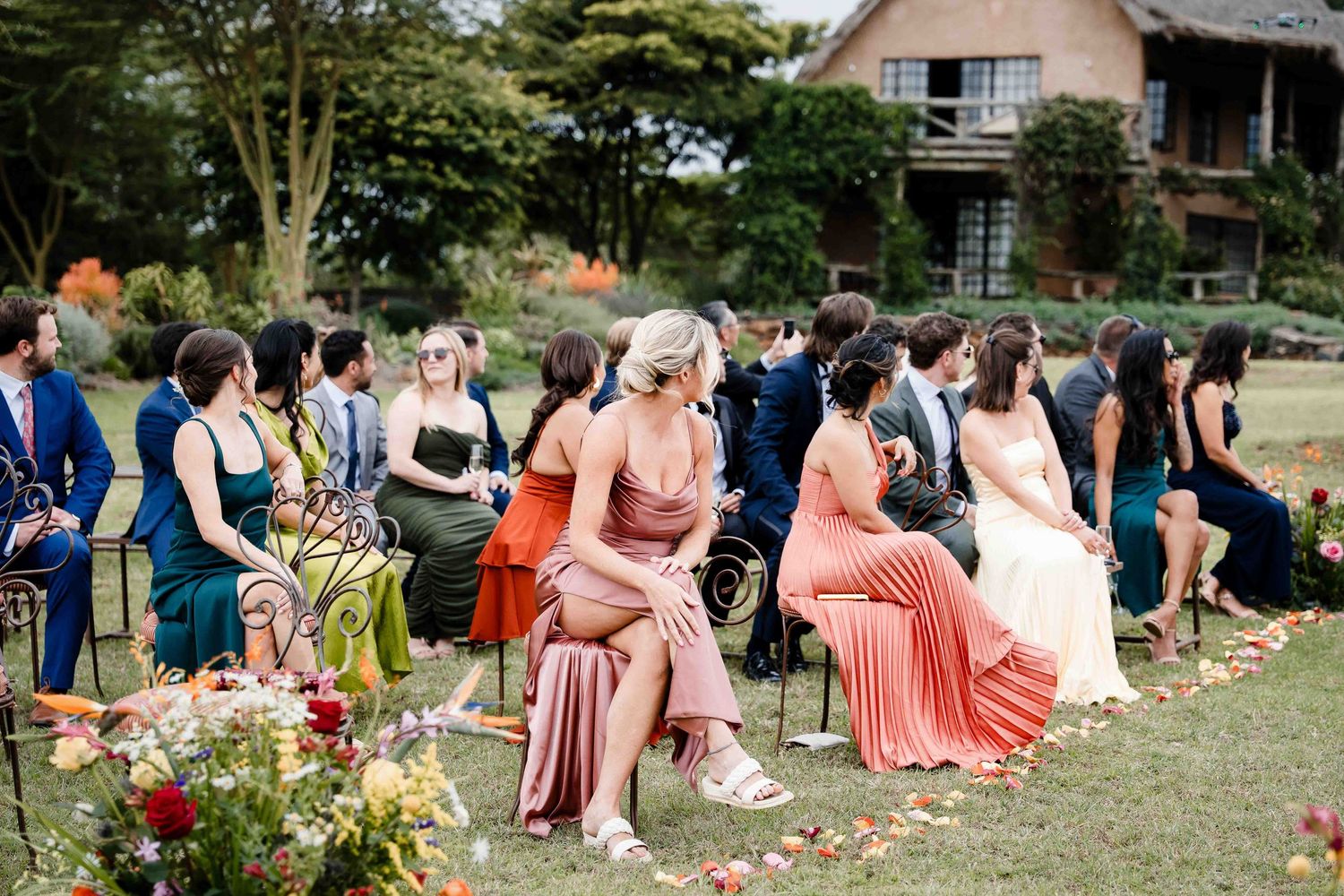Wedding guests seated in coral-colored dresses on white chairs during an outdoor garden ceremony.