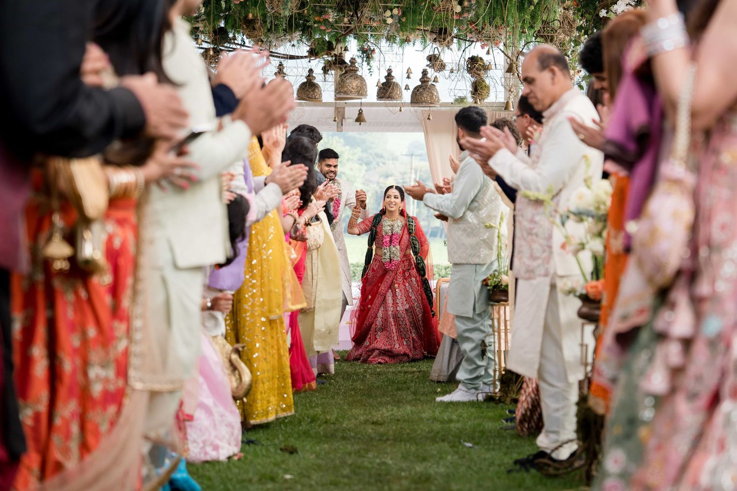 A traditional Indian wedding ceremony taking place outdoors under hanging floral decorations with guests lining both sides.