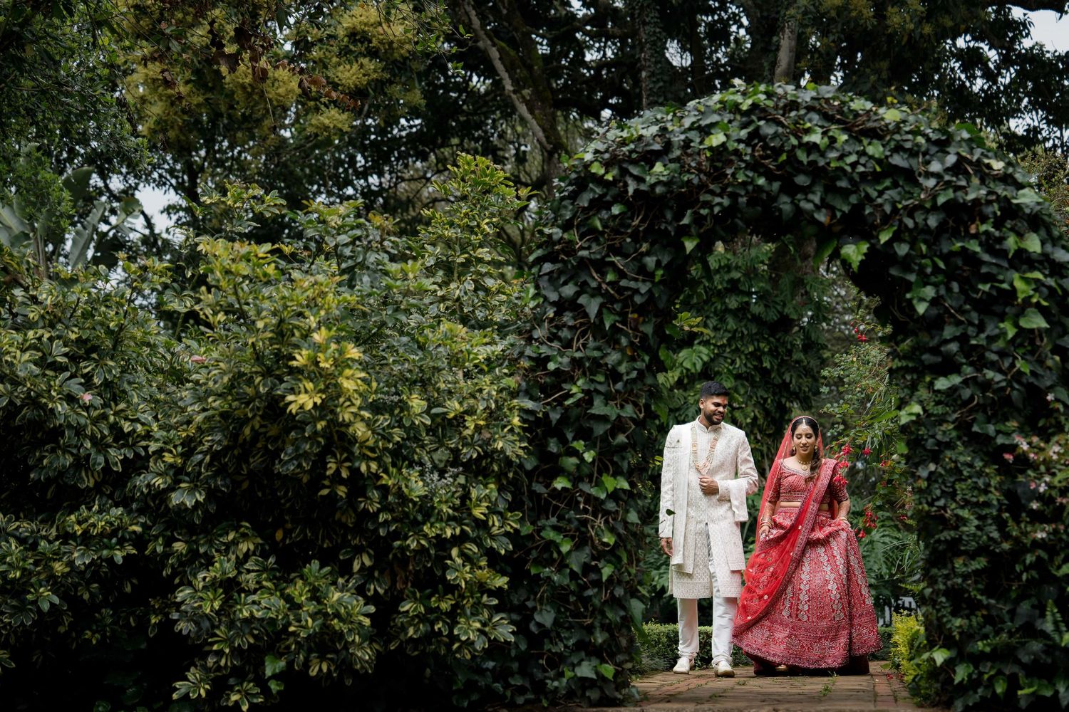 A couple in traditional wedding attire stands under a lush green garden archway during an outdoor ceremony.