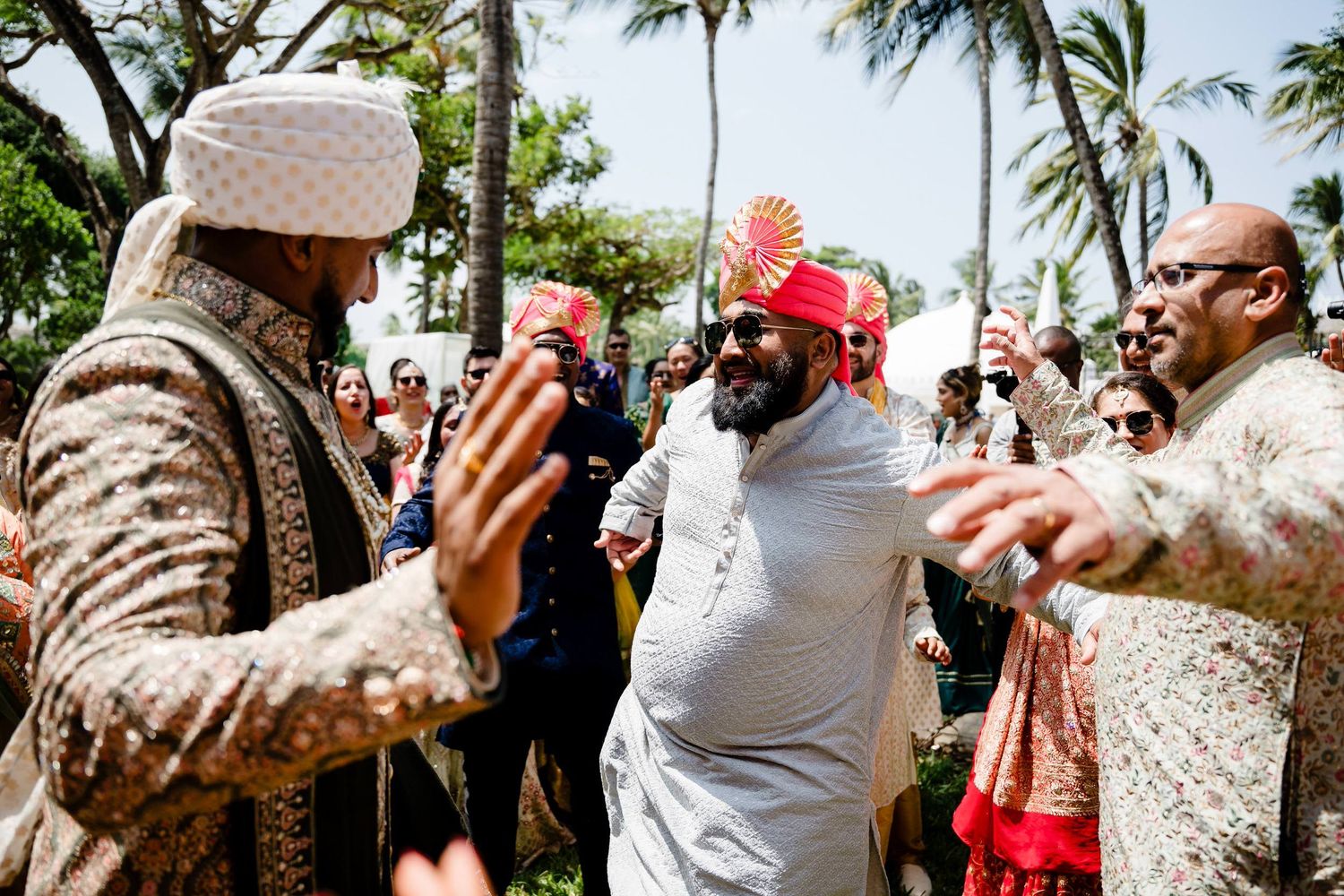 A group of people celebrating outdoors with drinks under palm trees at a festive daytime gathering.