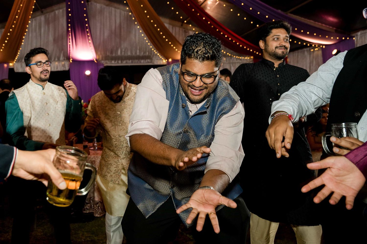Party guests dance and celebrate at an Indian wedding reception with string lights overhead.