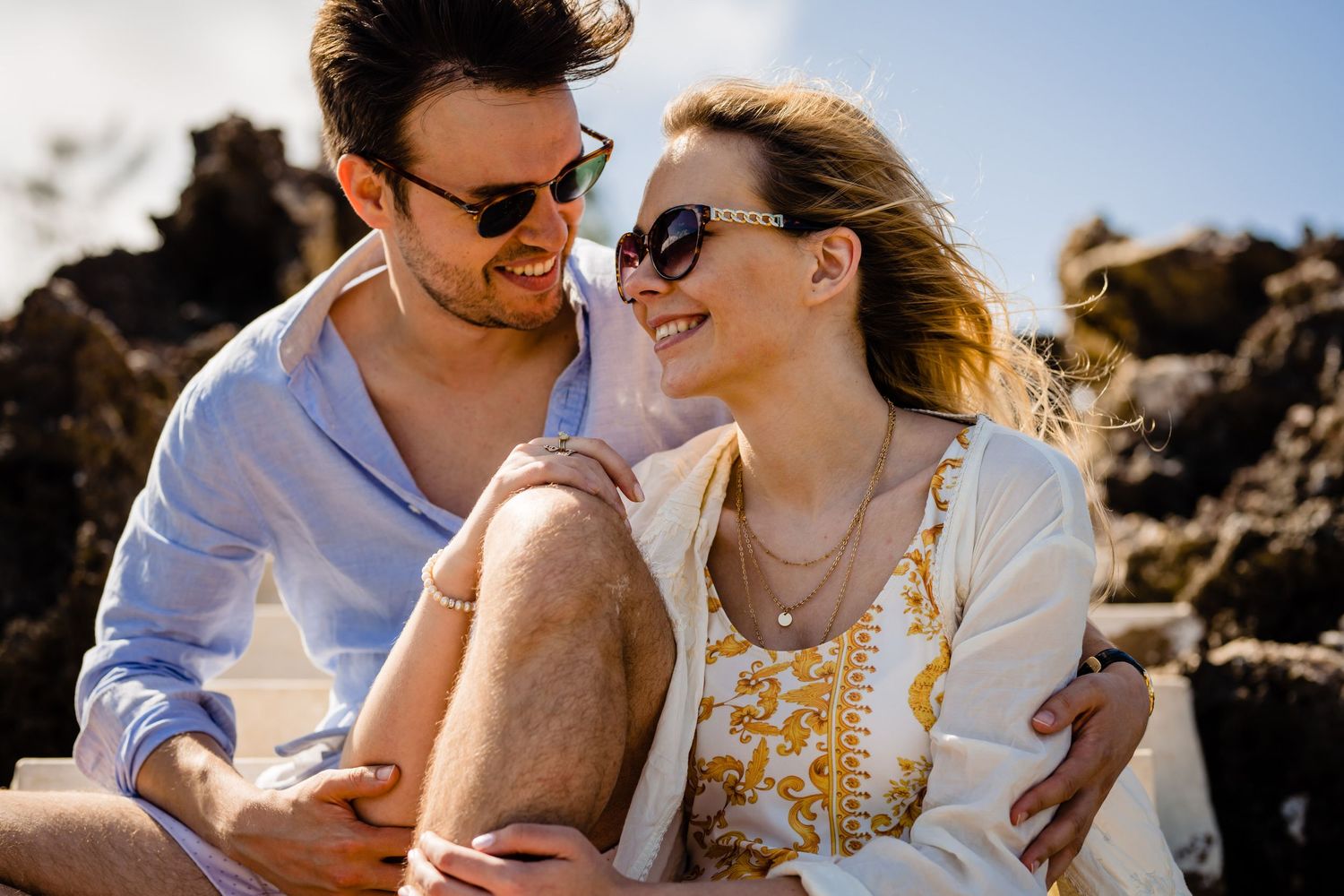 Joyful couples sharing tender moments on a sunny beach with ocean rocks in background.
