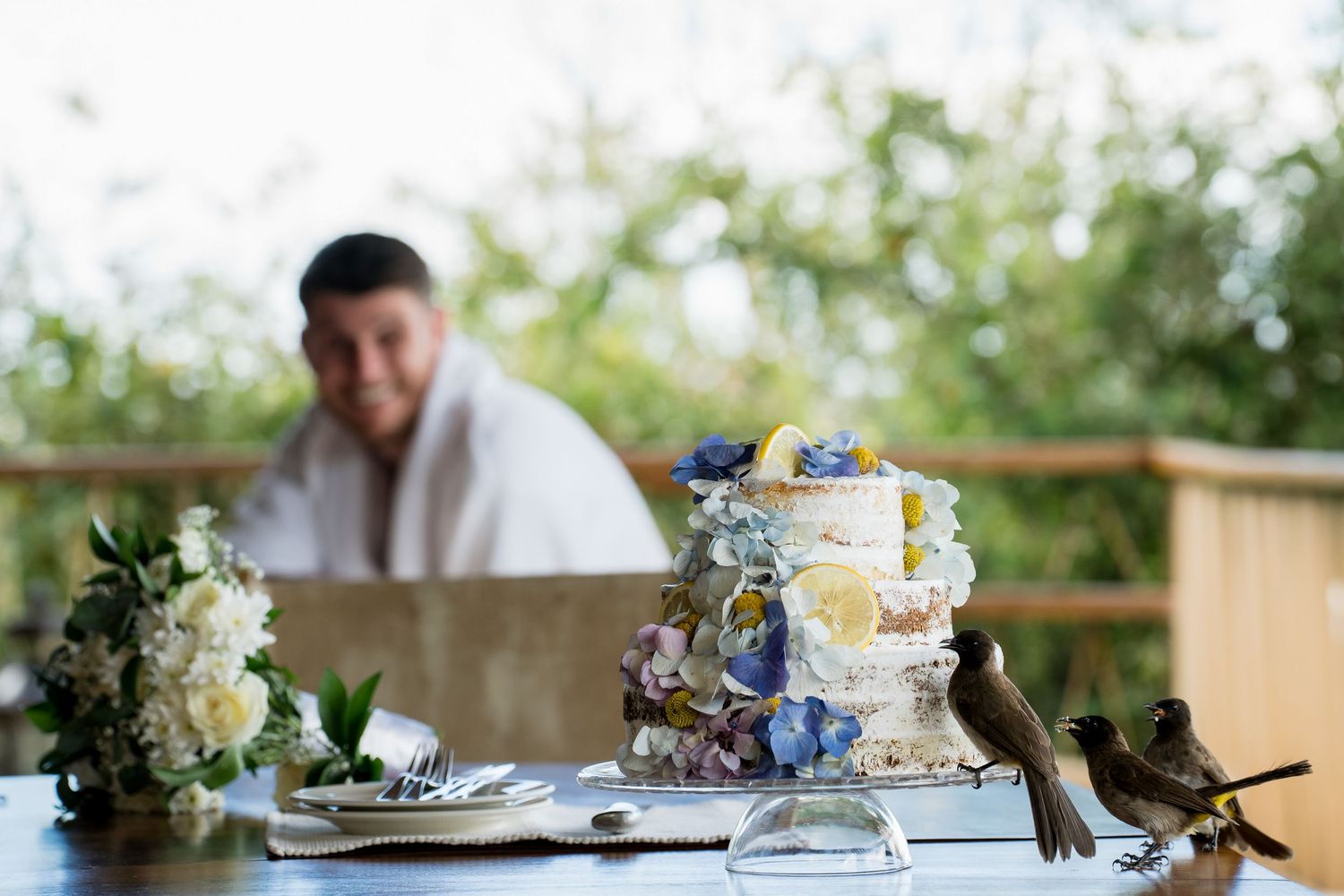 An elegant wedding cake with blue flowers sits on a decorated outdoor table.