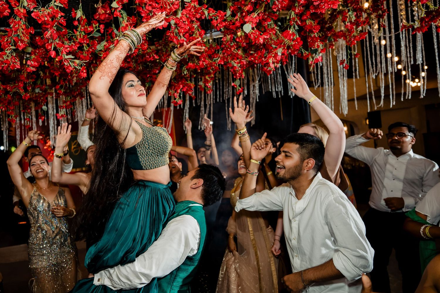 Guests dancing energetically at a reception under red floral ceiling decorations.