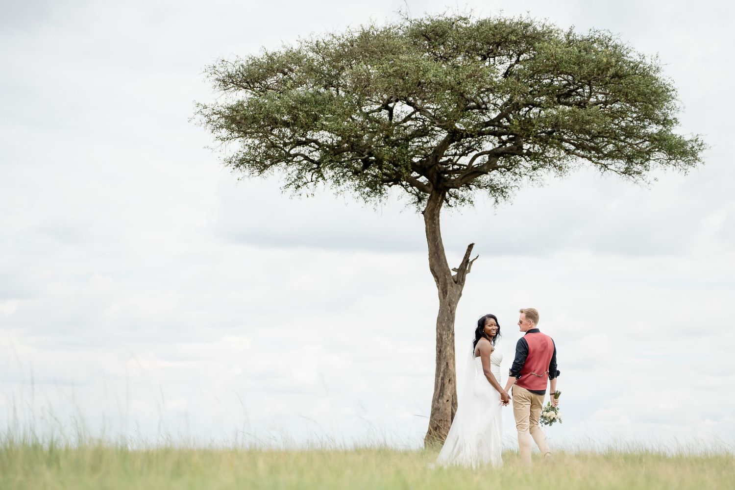 Wedding couple stands beneath lone acacia tree in African savanna landscape.