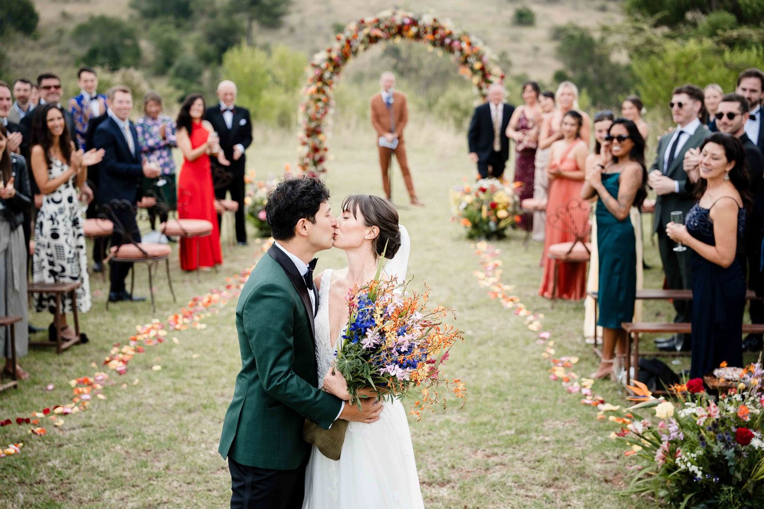 Wedding couple kisses at outdoor ceremony while guests look on from both sides of flower-lined aisle.