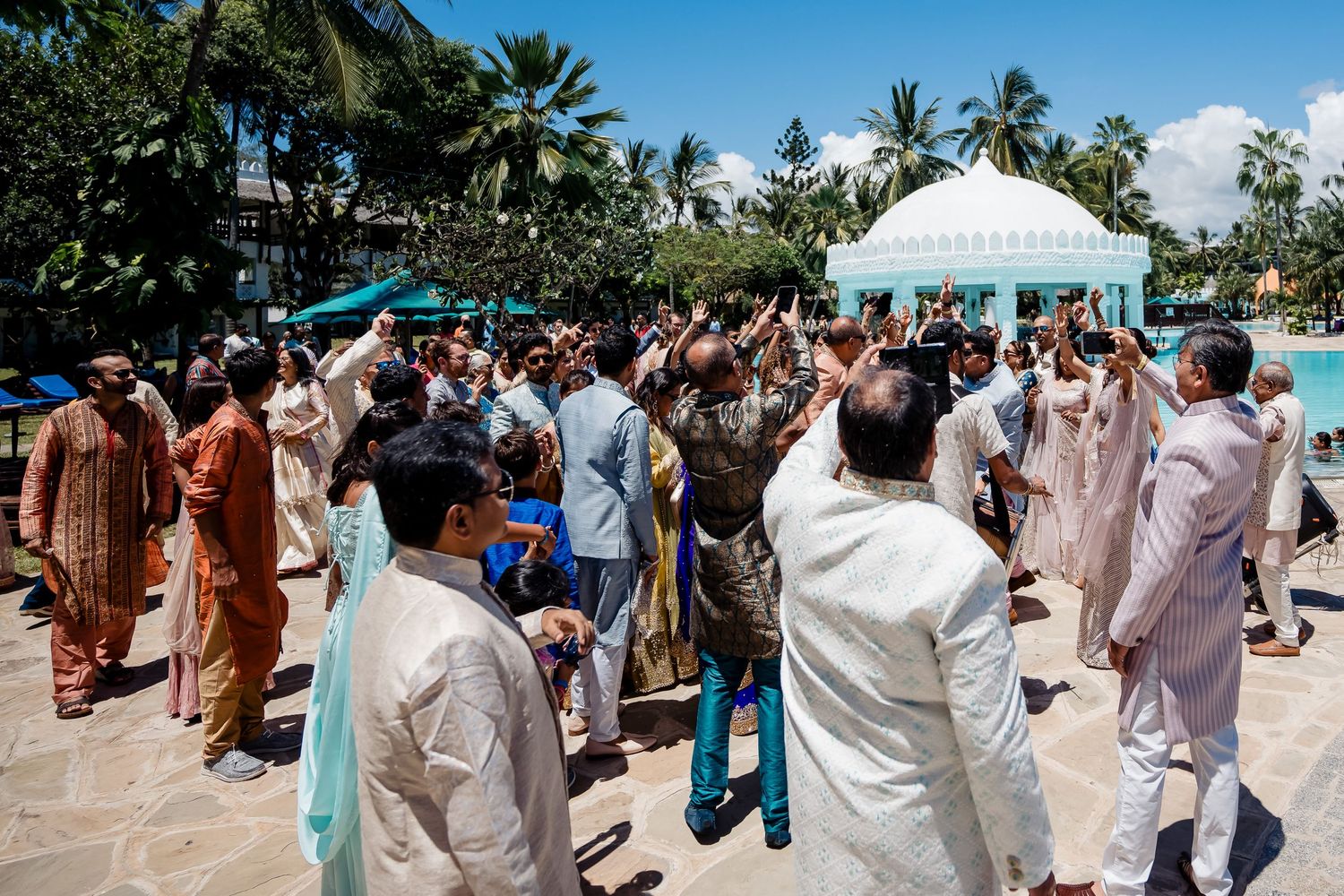 Guests in traditional cultural attire congregate under palm trees at an outdoor celebration.