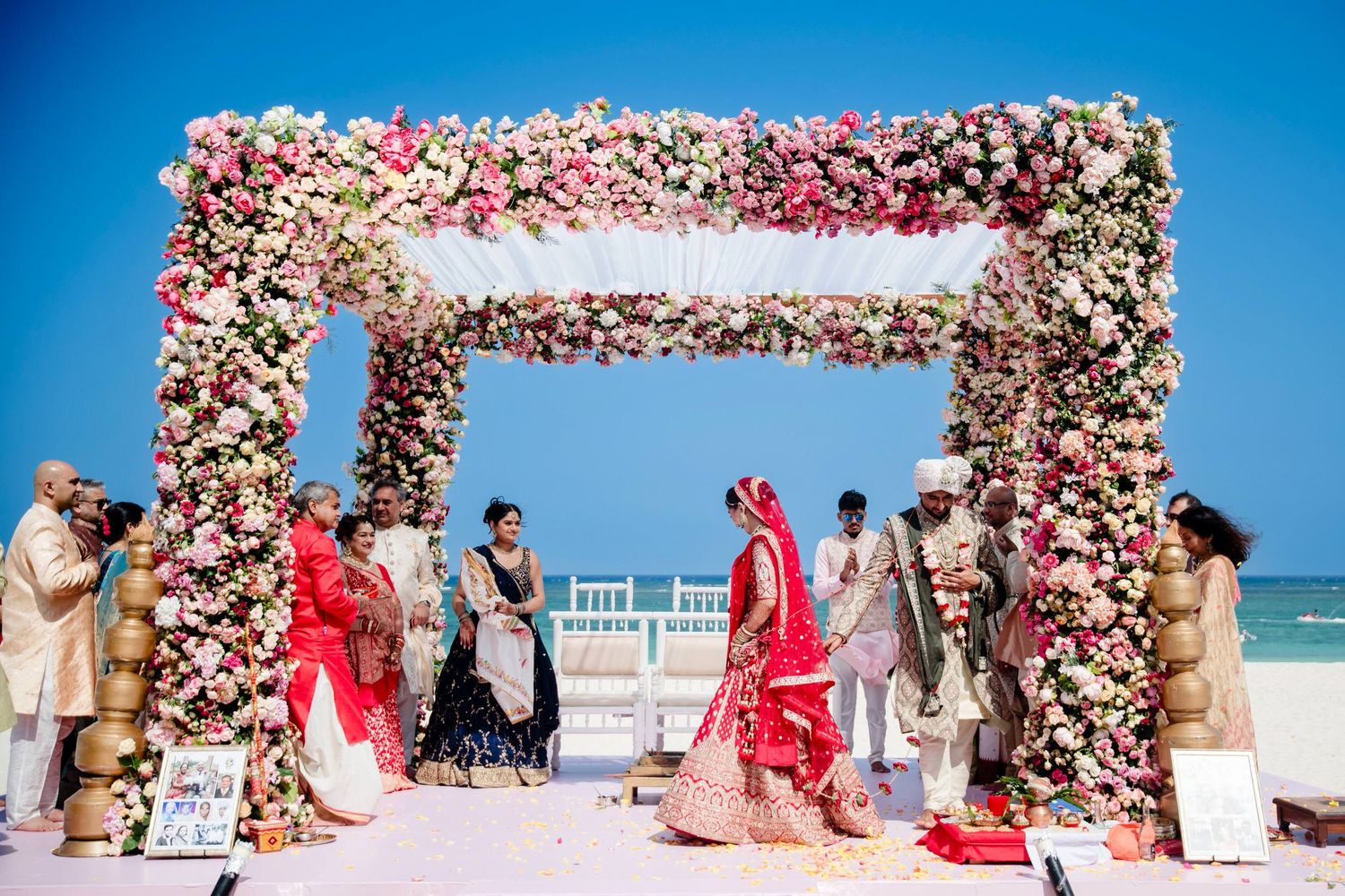 A floral arch with pink and white roses frames a beachside wedding ceremony setup against a bright blue sky.