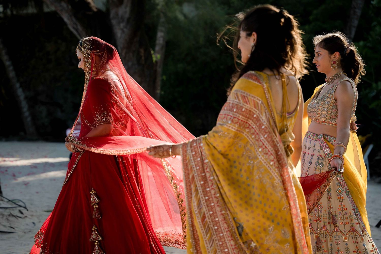 Traditional Indian wedding ceremony with bridesmaids wearing vibrant red and yellow lehengas walking in sunlight.