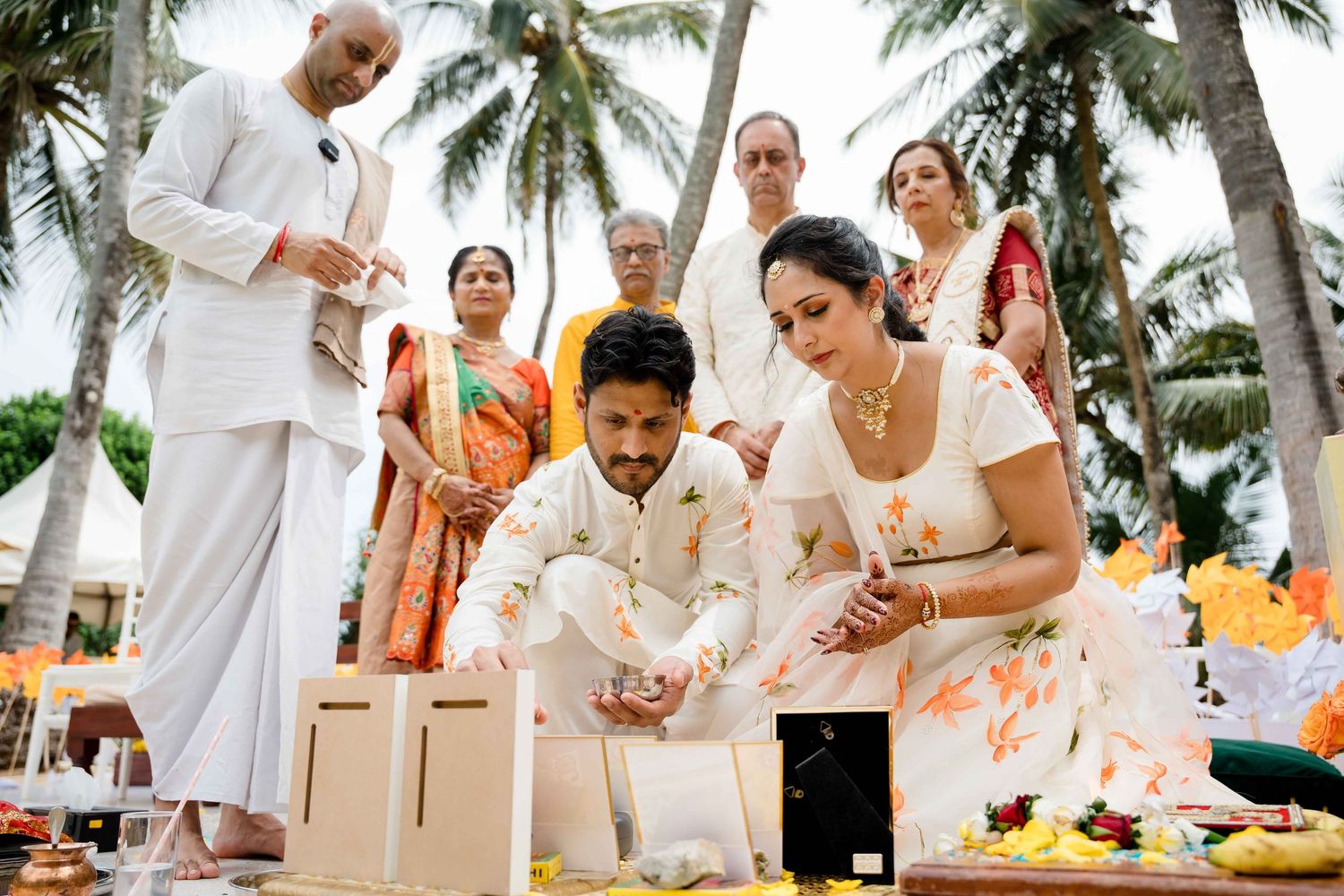 Tropical outdoor wedding ceremony setup with palm trees and white-clad guests gathered for a traditional ritual.
