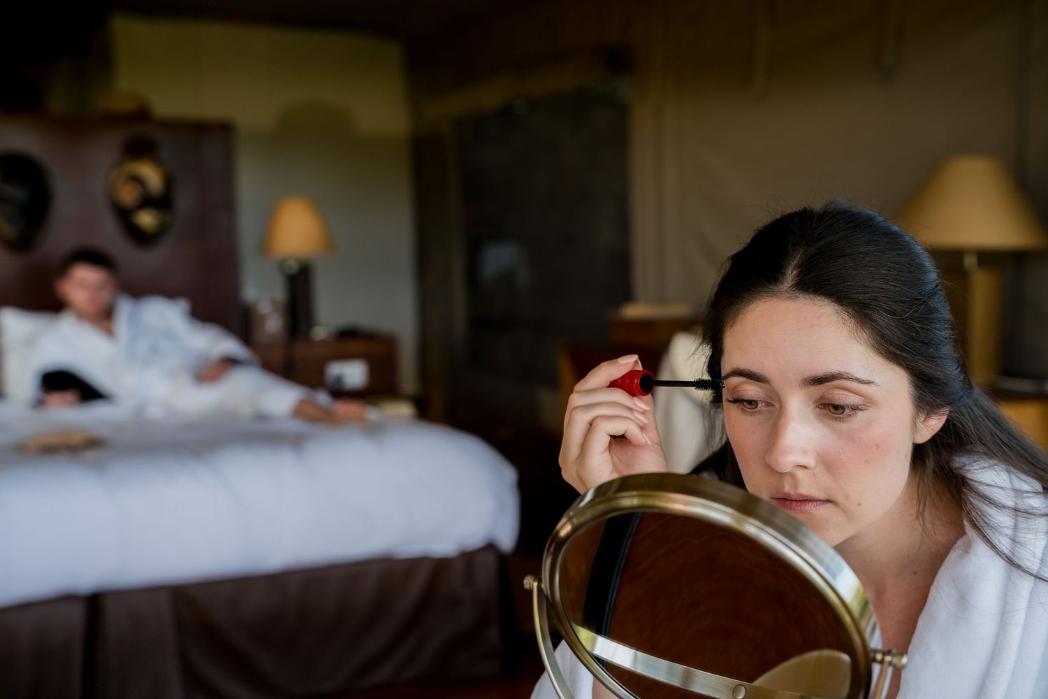 Woman in white robe applying mascara at vanity mirror in luxury hotel room with man resting on bed.