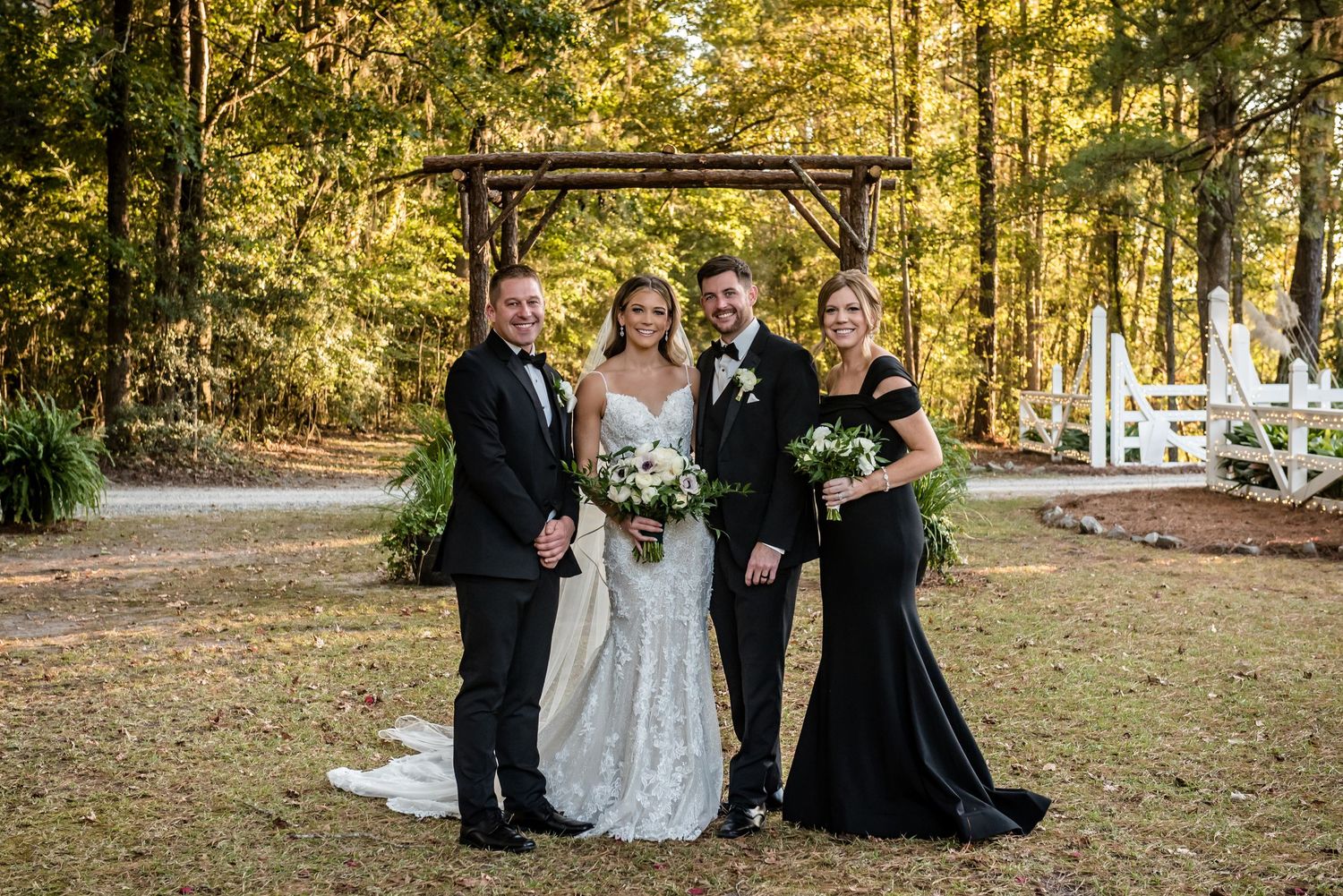 Wedding party of four posing outdoors in formal attire with rustic wooden arbor and white chairs among autumn trees.